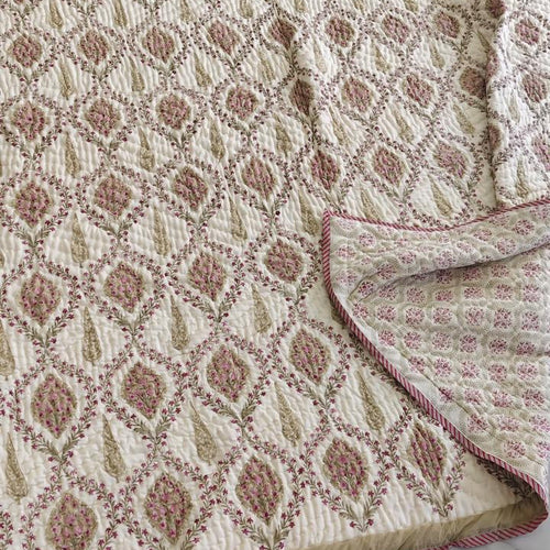 A woven throw blanket with a geometric diamond pattern in beige, cream, and muted pink, laid out on a light gray marble floor with one corner folded over.